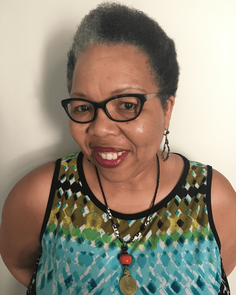 Portrait of a smiling woman with short natural hair and glasses wearing a patterned sleeveless top and necklace, photographed against a plain background.
